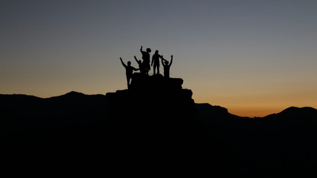 A silhouette of 5 people with arms raised up at the top of a mountain