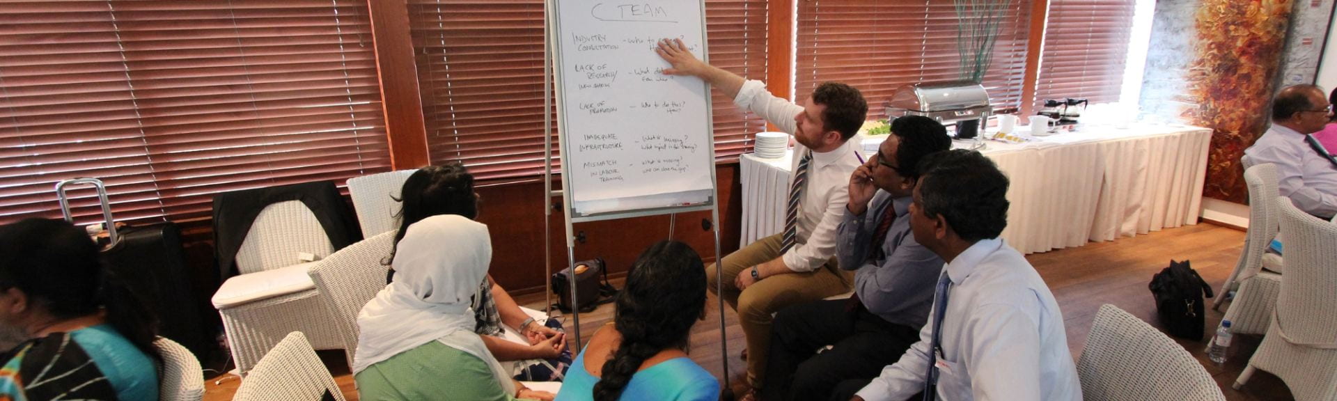 A group of people circled around a white board and pointing to the PDIA process on the whiteboard