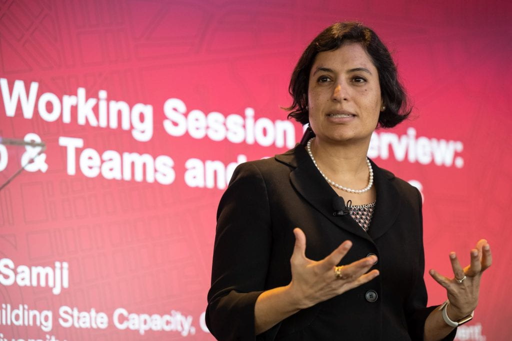 Salimah Samji wearing a black blazer and speaking in front of a red slide