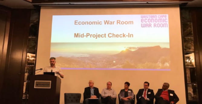 Presentation with slide title "Economic War Room: Mid-Project Check In" displayed behind presenter