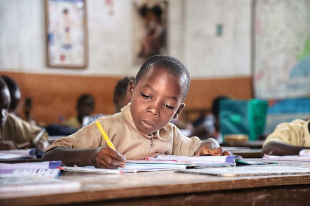 Young boy writing in a notebook at school desk with sudents in the background