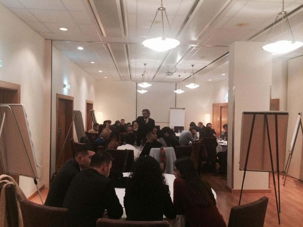 Matt Andrews speaking in a room with 30 audience members seated at circular tables and whiteboards throughout the room