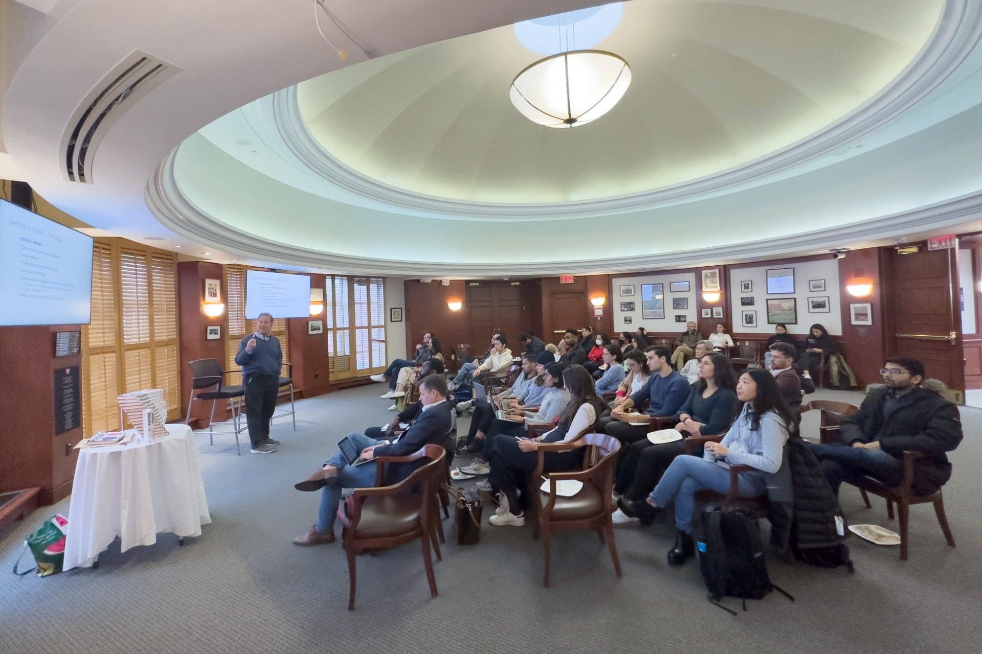Steve Kadish presenting in front of seated students in Allison dining room with his book stacked on a table and 2 screens with his presentation projected