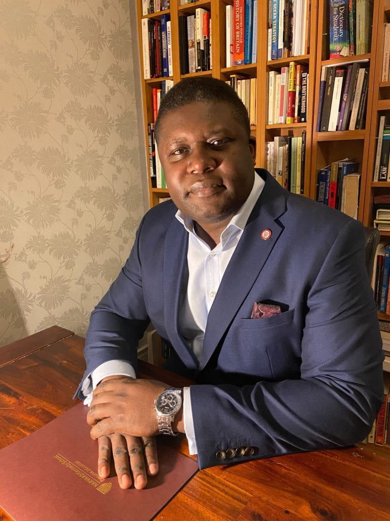 Bandi Mbubi in a blue blazer seated in front of a bookshelf