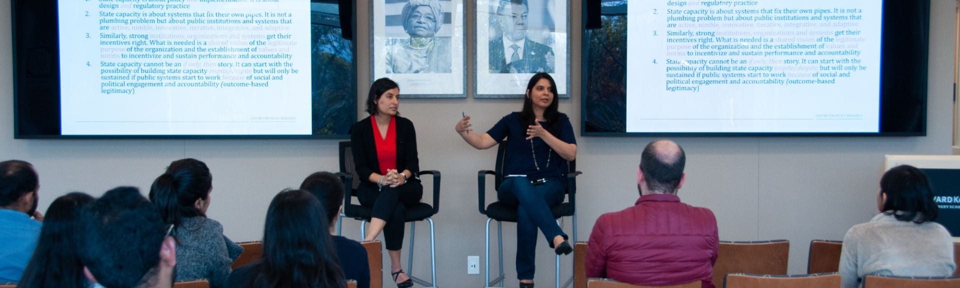 Two panelists seated in high chairs in the front of the room with screen projections behind them and audience members seated in front of them