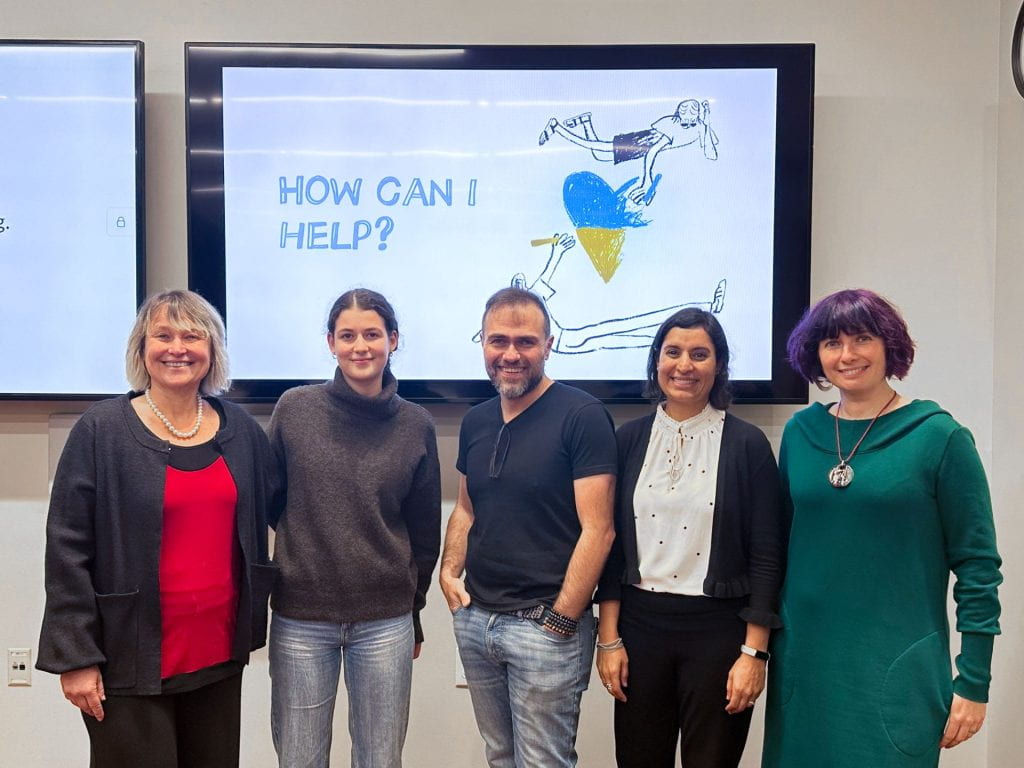 Panelists standing in front of a screen projecting "How can I help" with a heart filled with the Ukrainian flag colors