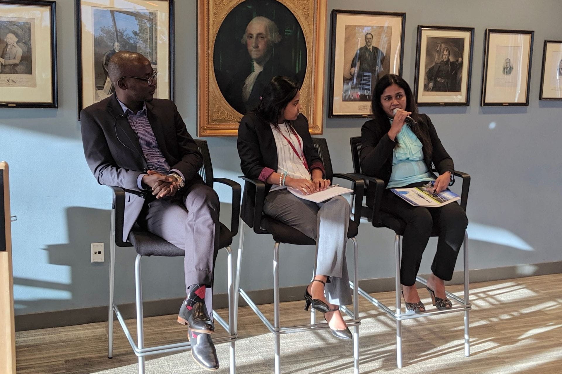 3 panelists seated in the front of the room with hanging portraits behind them