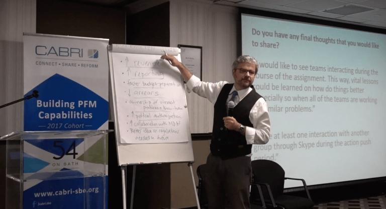 Matt Andrews writing on a whiteboard with a presentation slide behind him and CABRI PFM signage off to the side