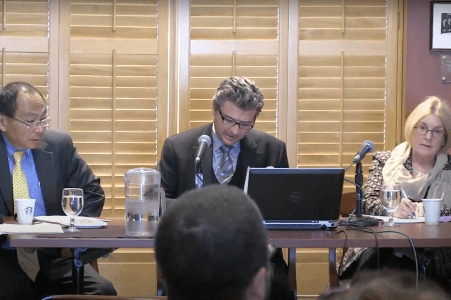 Matt Andrews sitting with two panelists and speaking in front of audience. A table and laptop is in front of them