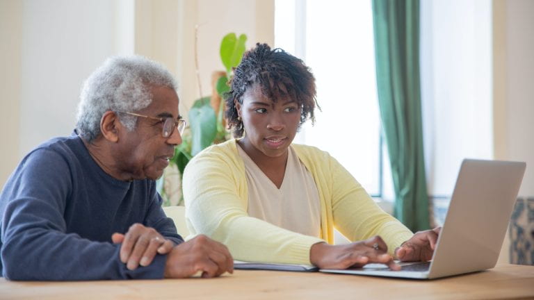 A woman using a laptop to assist and elderly man sitting next to her