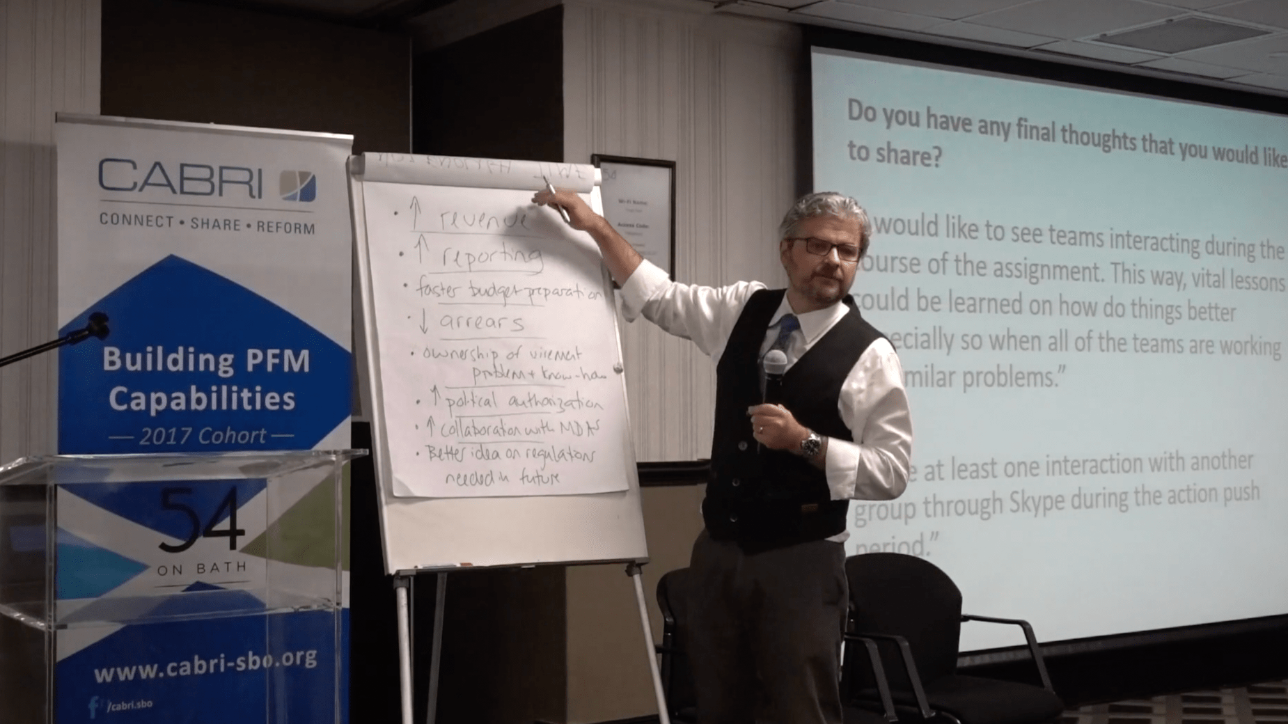 Matt Andrews writing on a whiteboard with a presentation slide behind him and CABRI PFM signage off to the side