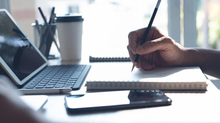 A person's hand writing notes with phone and laptop on table