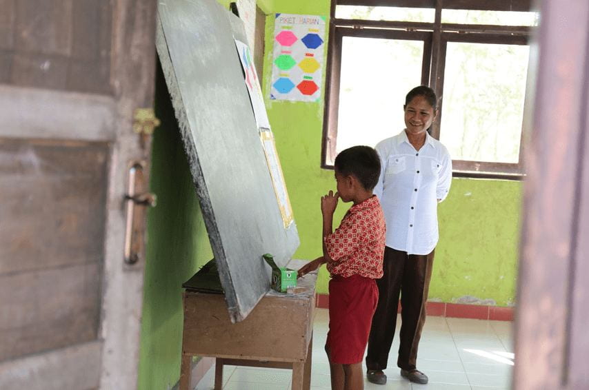 child writing on a black board as a woman supervises and laughs on