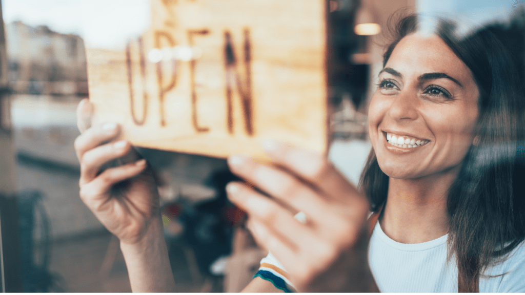 A woman stands behing an "Open" sign at her small business