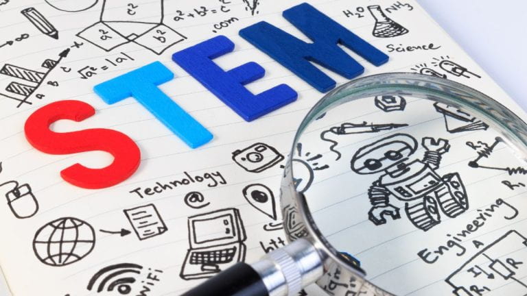 An image with STEM in block letters written on a notebook with a magnifying glass.