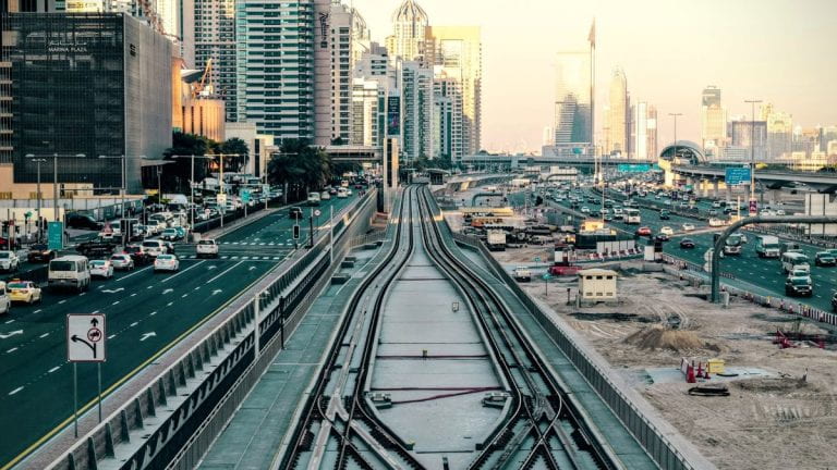 A cityscape with busy highways on both sides of an elevated metro track under construction, surrounded by tall buildings at sunset.