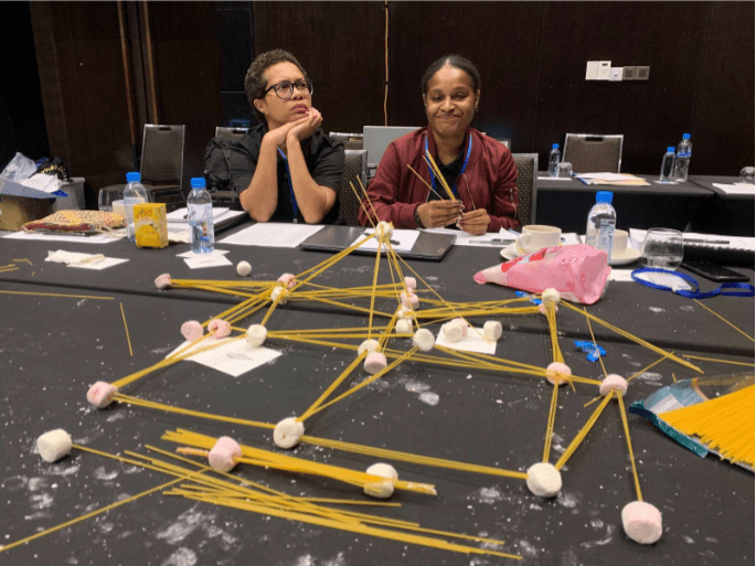 2 participants from Team Mobility Minds sitting at a table putting together a structure using spaghetti and marshmallows.