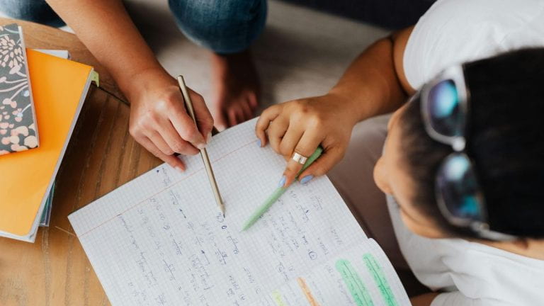 Overhead view of two people sitting at a desk and collaborating on handwritten notes in a grid-lined notebook, with one person pointing using a pen and the other holding a pencil; colorful notebooks are stacked nearby.