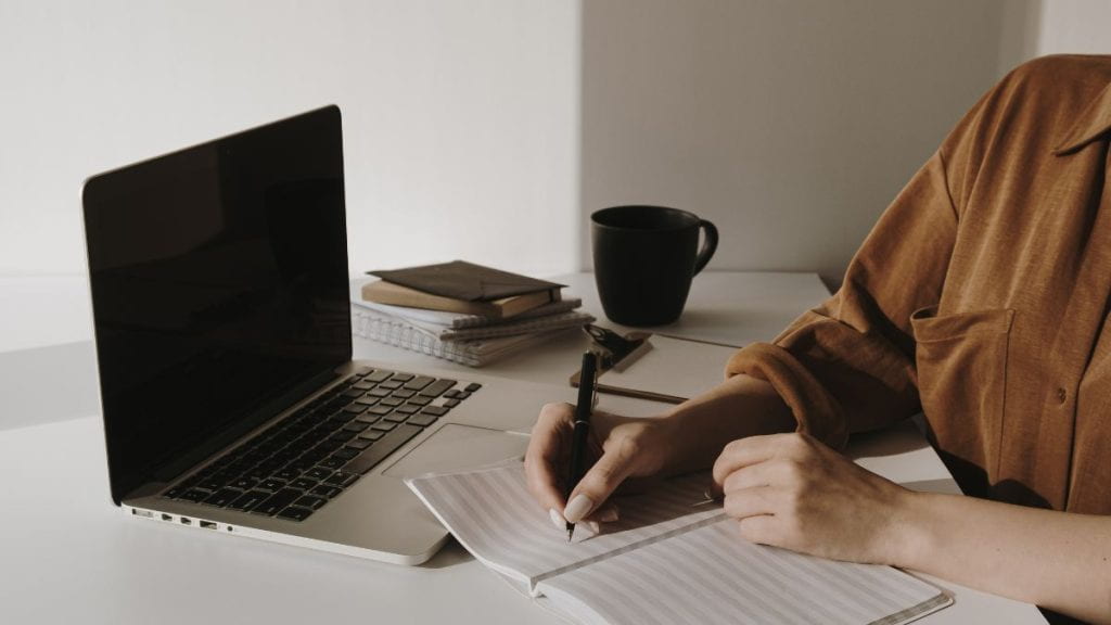 Person sitting at a desk writing in a notebook beside an open laptop, with stacked notebooks, and a black mug.